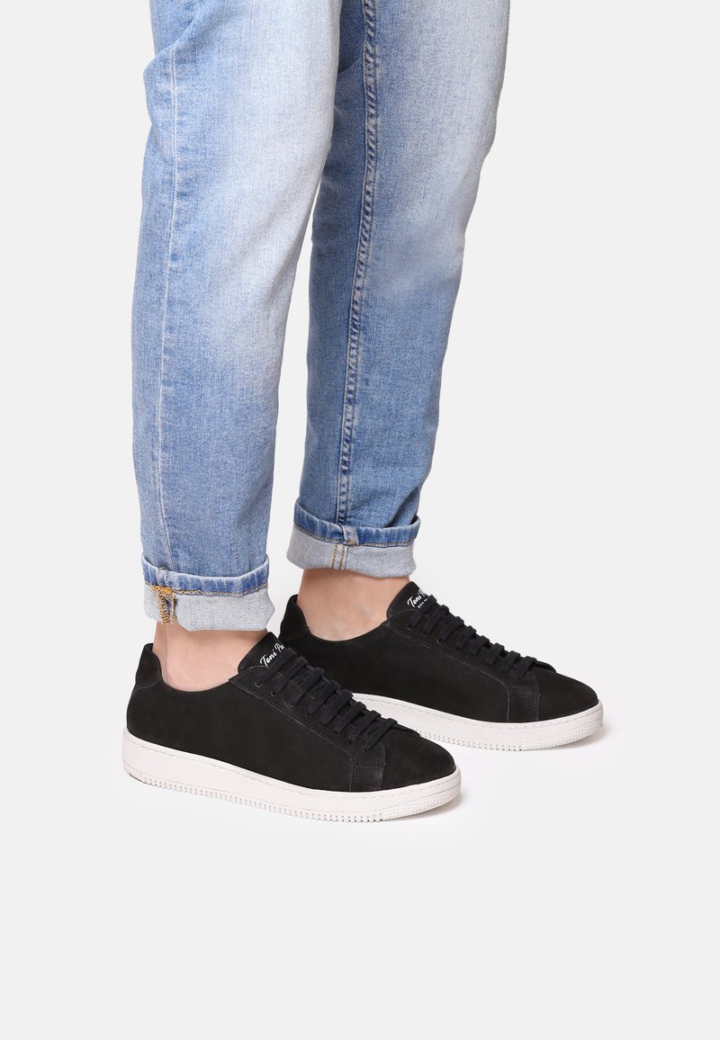 Zapatillas de ante negro con suelas de goma blanca, que cuentan con puntera redonda, diseño con cordones y un detalle minimalista. Combinadas con jeans de lavado claro con dobladillo.