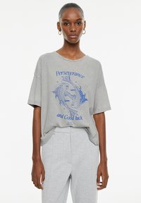 Camiseta gris de gran tamaño con mangas cortas, que presenta gráficos de peces azules y el texto "Perseverancia y buena suerte". Tejido texturizado.