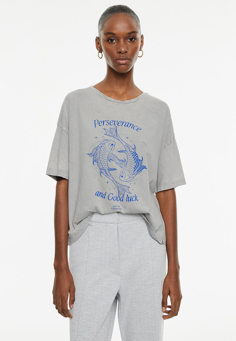 Camiseta gris de gran tamaño con mangas cortas, que presenta gráficos de peces azules y el texto "Perseverancia y buena suerte". Tejido texturizado.