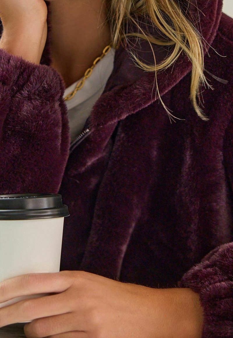 Veste zippée en peluche de couleur violet profond avec une texture douce ; tenant une tasse de café jetable avec un couvercle noir. Collier chaîne en or visible.