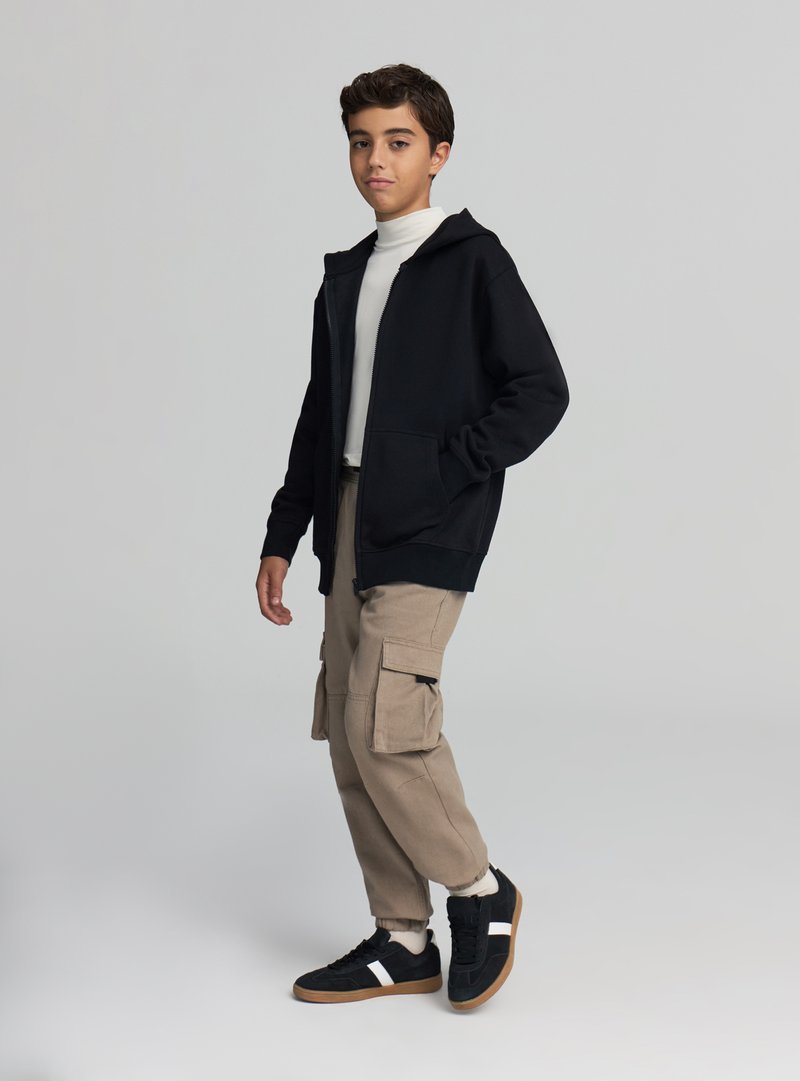 Felpa con cappuccio nera con zip sopra un dolcevita bianco, abbinata a pantaloni cargo beige e sneakers nere con strisce bianche. Abbigliamento casual.