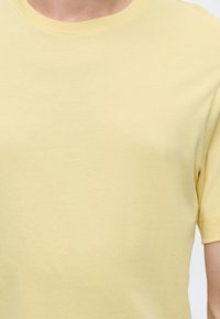 Personne portant un t-shirt jaune clair uni à manches courtes, montrant la partie supérieure de la poitrine et une partie du bras droit.