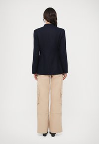 Tengerészkék blézer testhezálló szabásban, bézs cargo nadrággal, amelynek oldalzsebei vannak, magas derékkal és széles, harangszárú lábakkal. Fekete magassarkú cipővel egészül ki az öltözet.