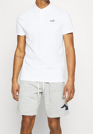 Man draagt een wit poloshirt met korte mouwen en lichtgrijze short met trekkoord en een donker logo op het linkerbeen.