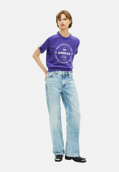 Młoda osoba stojąca z rękami za plecami, ubrana w fioletowy sweter z grafiką, jasnoniebieskie szerokie dżinsy z ozdobami oraz czarne buty.