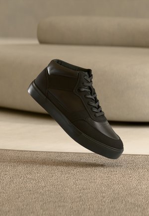 Czarny wysoki sneaker ze sznurowadłami unoszący się nad beżowym dywanem w pomieszczeniu o neutralnych tonach z sofą w tle.
