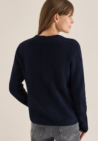 Dunkelblauer gestrickter Pullover mit gerippter Textur, langen Ärmeln und einem Rundhalsausschnitt. Hat einen leichten Abfall am hinteren Saum und minimale Akzente.