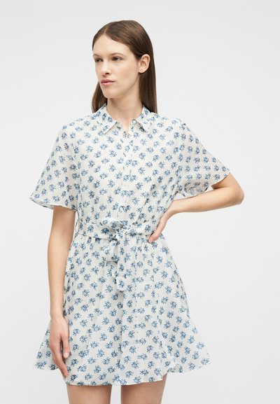 Γυναίκα που φοράει λευκό φόρεμα με κοντά μανίκια, μπλε λουλουδάτο μοτίβο και ζώνη στη μέση, στέκεται με το ένα χέρι στη μέση μπροστά σε απλό φόντο.