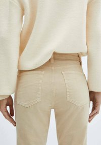 Beige high-waisted jeans met een gestructureerde afwerking, voorzien van twee achterzakken en subtiele stikseldetails. De hand van het model rust op de heup.