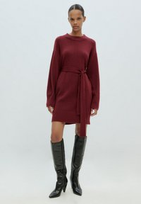 Bordeauxfarbenes Strickkleid mit Rundhalsausschnitt und tailliertem Gürtel. Lange Ärmel, gerippte Bündchen und kurzer Saum. Das Modell trägt kniehohe schwarze Stiefel mit Absatz.