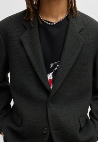 Blazer scuro e strutturato con rever a scollo, due tasche frontali e due bottoni. Indossato sopra una t-shirt nera con grafica.