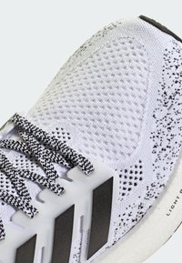 Białe buty sportowe z teksturowaną cholewką z dzianiny, czarnymi i białymi sznurowadłami oraz trzema czarnymi paskami po bokach. Cechuje się elastycznym designem podeszwy.