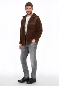 Chaqueta de forro polar marrón con una sección superior marrón, cierre frontal con cremallera y capucha. Se lleva puesta sobre una camisa blanca con jeans grises y botas negras.