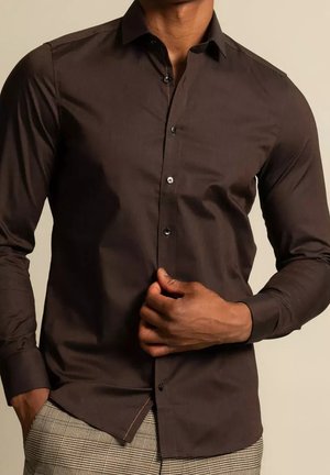 Mężczyzna zapinający ciemnobrązową koszulę z długim rękawem, ubrany w szare spodnie w kratę, stojący na jednolitym beżowym tle.