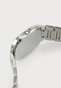 Hõbedane roostevabast terasest kell ümmarguse korpuse ja tasase klaasiga, millel on sile bezel ja seotud käevõru. Näo detaile pole nähtav.