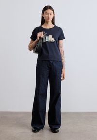 Μπλε σκούρο βαμβακερό μπλουζάκι με γραφικό τύπωμα ενός σκύλου και λουλουδιών, συνδυασμένο με σκούρο τζιν παντελόνι φαρδιάς γραμμής και μαύρη τσάντα ώμου.