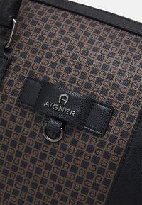 Sac Aigner avec une texture en brun et noir à motifs. Il dispose d'une bandoulière en cuir noir et d'un logo argenté sur un onglet rectangulaire.