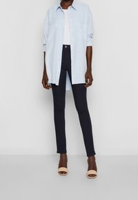 Světle modrá oversized košile s detaily knoflíků, kombinovaná s tmavými skinny džíny a nahými sandály na vysokém podpatku. Hladká tkanina a minimalistický design.