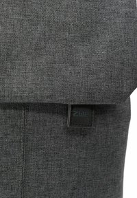 Mochila de tela gris con un acabado texturizado, que presenta una parte superior enrollada, una etiqueta de logo negra y detalles de costura visibles.