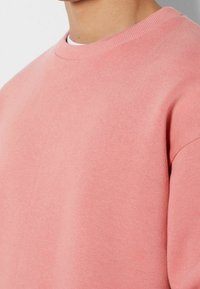 Närbild av en person som bär en enkel rosa crewneck sweatshirt över en vit undertröja, där axel- och övre bröstdetaljer visas.