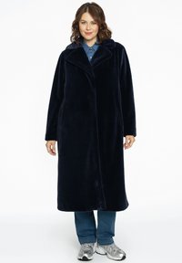 Marineblå faux pelsfrakke med krave, lange ærmer og en lige silhuet, kombineret med denimjeans og sølvfarvede sneakers.