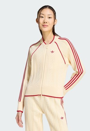 Vrouw draagt roomkleurig gebreid Adidas trainingspak met rode strepen en logo's, staand tegen een effen lichte achtergrond met hand op heup.