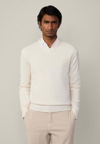 Cremefarbener V-Ausschnitt-Pullover aus weichem Strickmaterial, kombiniert mit einem weißen Hemd mit Kragen. Verfügt über gerippte Bündchen und einen gerippten Saum, dezentes Branding sichtbar.