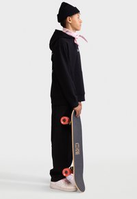 Juodas džemperis su šoninėmis angomis, šviesiai rožinė kaklo skara, juodos kelnės ir baltos sportbačiai. Laiko riedlentę su raudonais ratukais ir mediniu stogu.