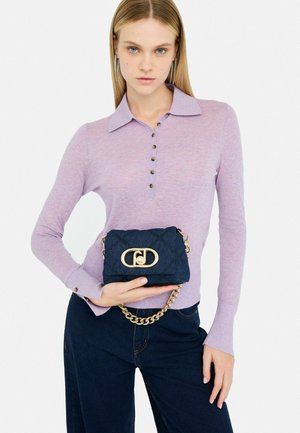Borsa in tessuto trapuntato di colore blu navy con dettagli in hardware dorato e tracolla a catena, tenuta con una mano. Il modello indossa una camicia leggera di colore viola chiaro con colletto.