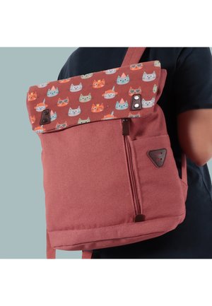 Rucksack mit gerolltem Oberteil, das bunte Katzenköpfe zeigt, getragen von einer Person in dunklem Shirt, vor einem einfarbigen hellblauen Hintergrund.