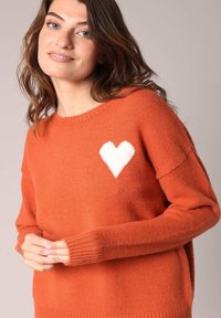 Pull orange en tricot doux. Présente un cœur blanc sur la poitrine, des poignets côtelés et un col rond classique.