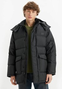 Giacca puffer nera con cappuccio, dotata di chiusura a zip frontale, due tasche anteriori e una finitura texturizzata. Indossata sopra una camicia verde.