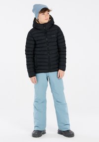 Chaqueta de plumas negra con capucha, con costuras horizontales. Pantalones azul claro, de tela impermeable, y botas de goma negras, de pie sobre un fondo blanco.
