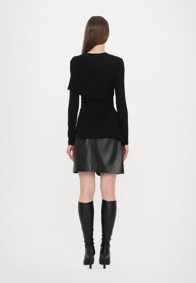 Schwarzes Langarmoberteil mit drapierter Design, kombiniert mit einem glänzenden schwarzen Lederrock. Das Modell trägt knielange schwarze Stiefel, die eine elegante Silhouette betonen.