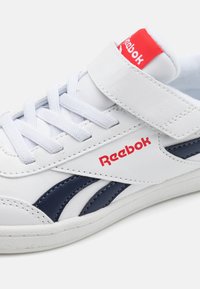 Zapatillas blancas de cuero, con un acento en azul marino, marca Reebok en rojo y una correa de enganche junto a los cordones tradicionales.