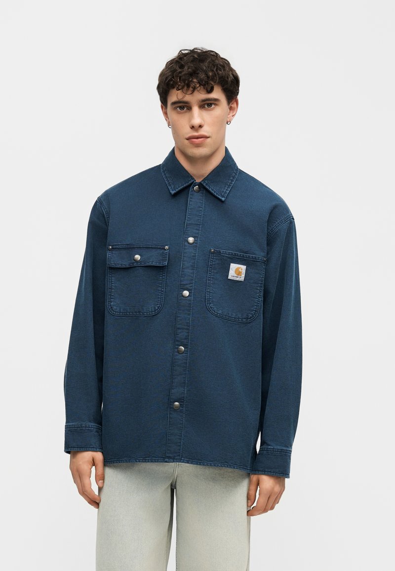 Junger Mann trägt eine dunkelblaue Jeansjacke mit Knöpfen und zwei Brusttaschen, eine mit einem Logo-Aufnäher, und eine hellbeige Hose vor einfachem Hintergrund.