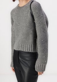 Donna che indossa un maglione grigio a maglia con piccoli dettagli e pantaloni di pelle neri, mentre porta una borsa a tracolla nera.