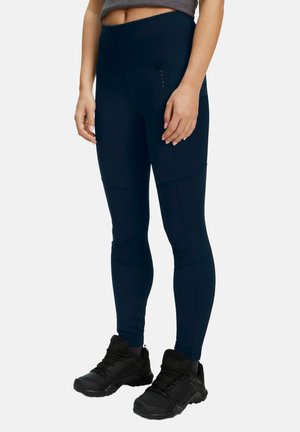 Femme portant des leggings bleu marine avec poches latérales et des chaussures de randonnée noires, debout devant un fond uni.