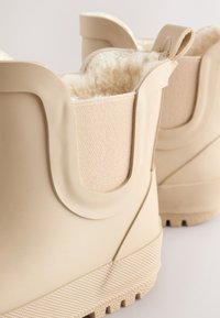 Bottes imperméables beiges montant à la cheville avec panneaux latéraux élastiques et doublure en polaire, montrées en gros plan.