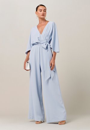 Žena v svetlomodrom overal s širokými nohavicami, opaskom na viazanie v páse a výstrihom do V, drží bielu kabelku a stojí na neutrálnej farbe pozadia.