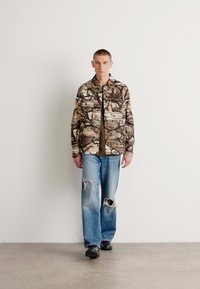 Camouflage-patterned shirt jacket with a collar, paired with light blue distressed jeans. Black shoes and a plain olive undershirt.