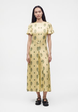 Femme aux longs cheveux foncés portant une robe midi jaune avec des motifs floraux verts et des chaussures Mary Jane noires, debout sur un fond blanc.