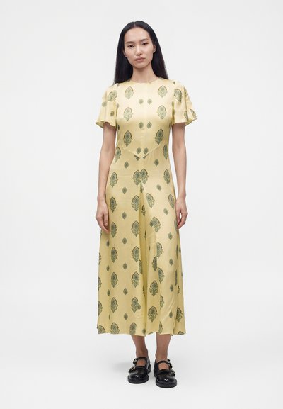 Femme aux longs cheveux foncés portant une robe midi jaune avec des motifs floraux verts et des chaussures Mary Jane noires, debout sur un fond blanc.