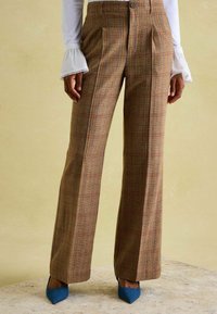 Rjave karo kariraste hlače z visokim pasom in širokimi hlačnicami, z gumbno zapenjanjem in ostrimi pregibi. Kombinirane z modrimi čevlji z ostro konico.