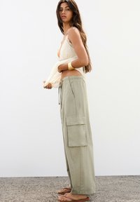 Lichtbeige cami met kantafwerking, gecombineerd met losse, lichtgroene cargobroek met twee zijzakken. Lang bruin haar, platte sandalen.