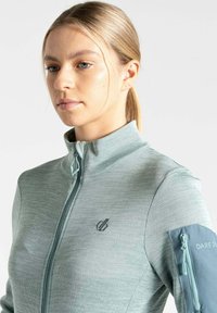 Hellgrüne Zip-Jacke mit strukturiertem Finish, hohem Kragen und kontrastierender blauer Packtasche mit Reißverschlüssen an den Ärmeln. Mit einem Logo auf der Brust.