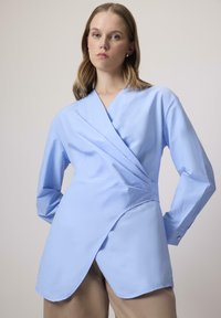 Világoskék, hosszú ujjú, kötött blúz, strukturált dizájnnal és ívelt hempergővel. Simogatós anyagból készült, amely finom textúrával és gombos díszítéssel rendelkezik.