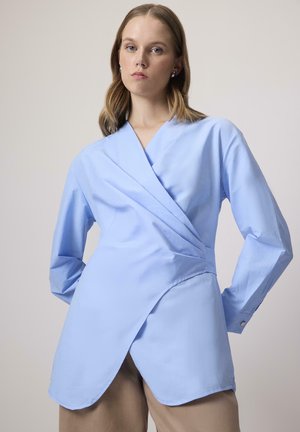 Lyseblå indpakningsbluse med lange ærmer, struktureret design og buet kant. Lavet af glat stof med subtil tekstur og knapdetaljer.