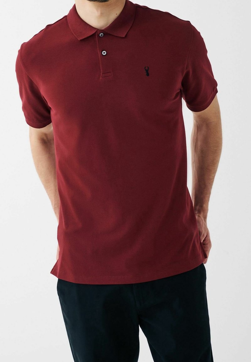 Polo en bourgogne fabriqué en coton, avec un col, un plastron à trois boutons, des manches courtes et un petit détail de logo noir.