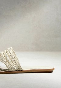 Sandale plate avec bride en cuir tressé blanc cassé et semelle couleur tan sur une surface et un fond gris clair.